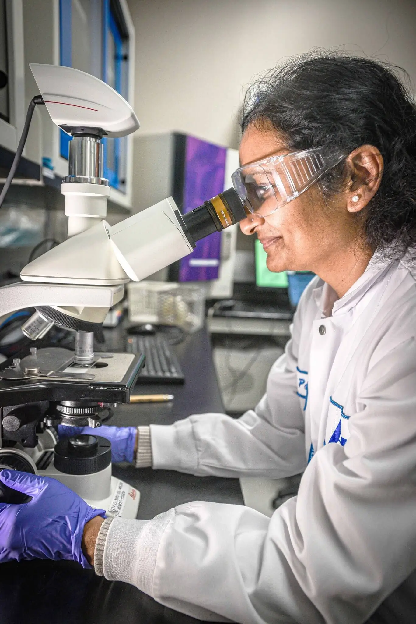 A scientist in a lab coat and safety goggles using a microscope.