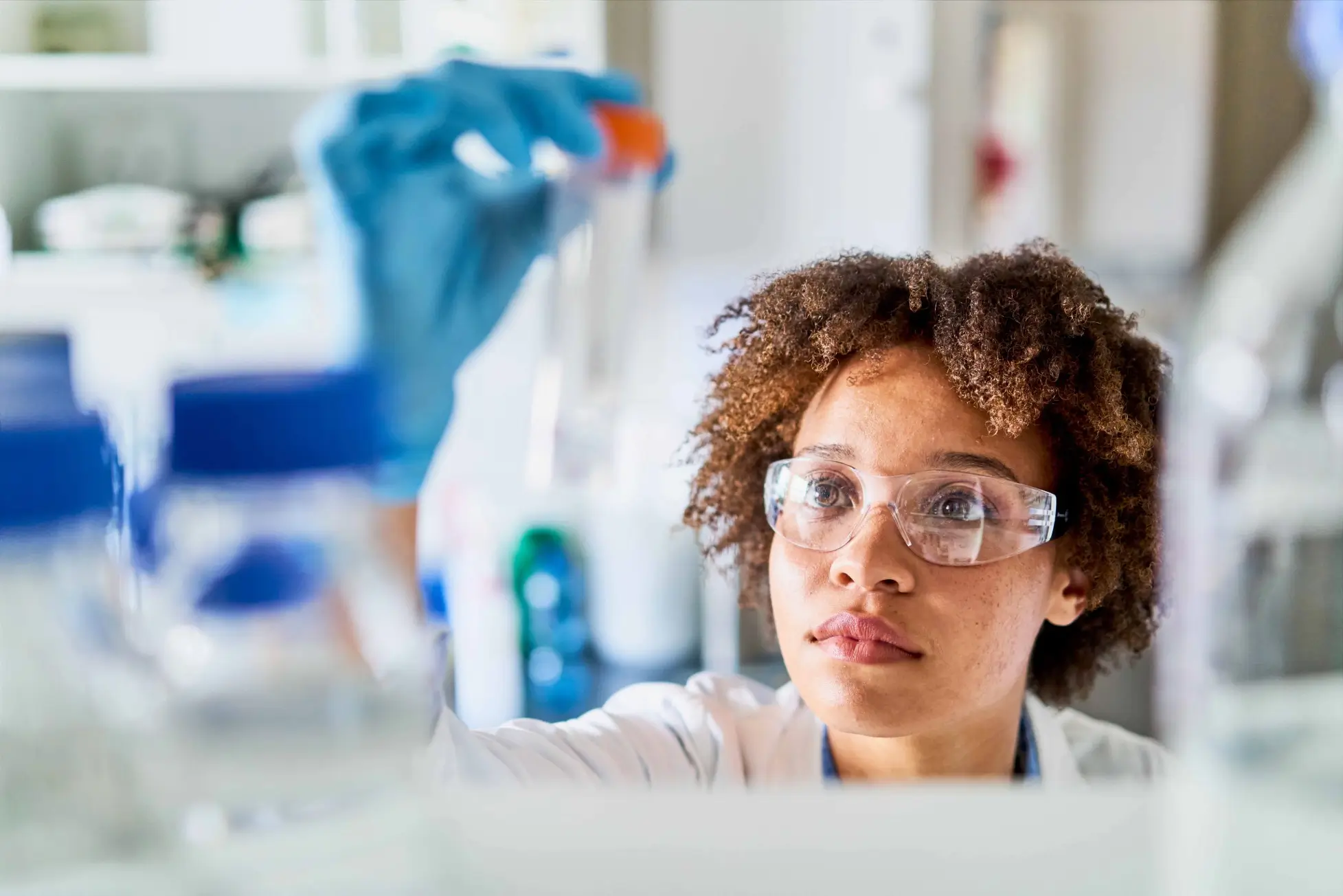 A woman in a lab coat and safety goggles holds a flask with a gloved hand