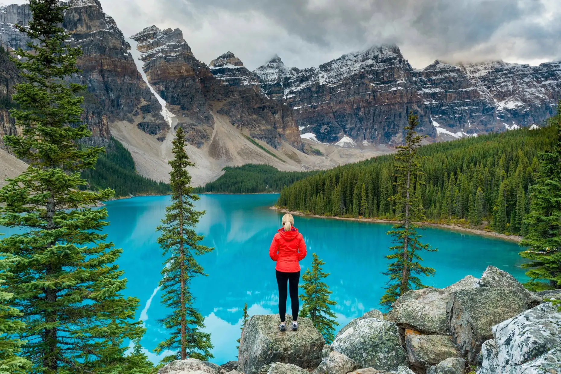 Une personne portant une veste rouge se tient sur des rochers surplombant un lac turquoise entouré d'arbres à feuilles persistantes et de montagnes enneigées sous un ciel nuageux.