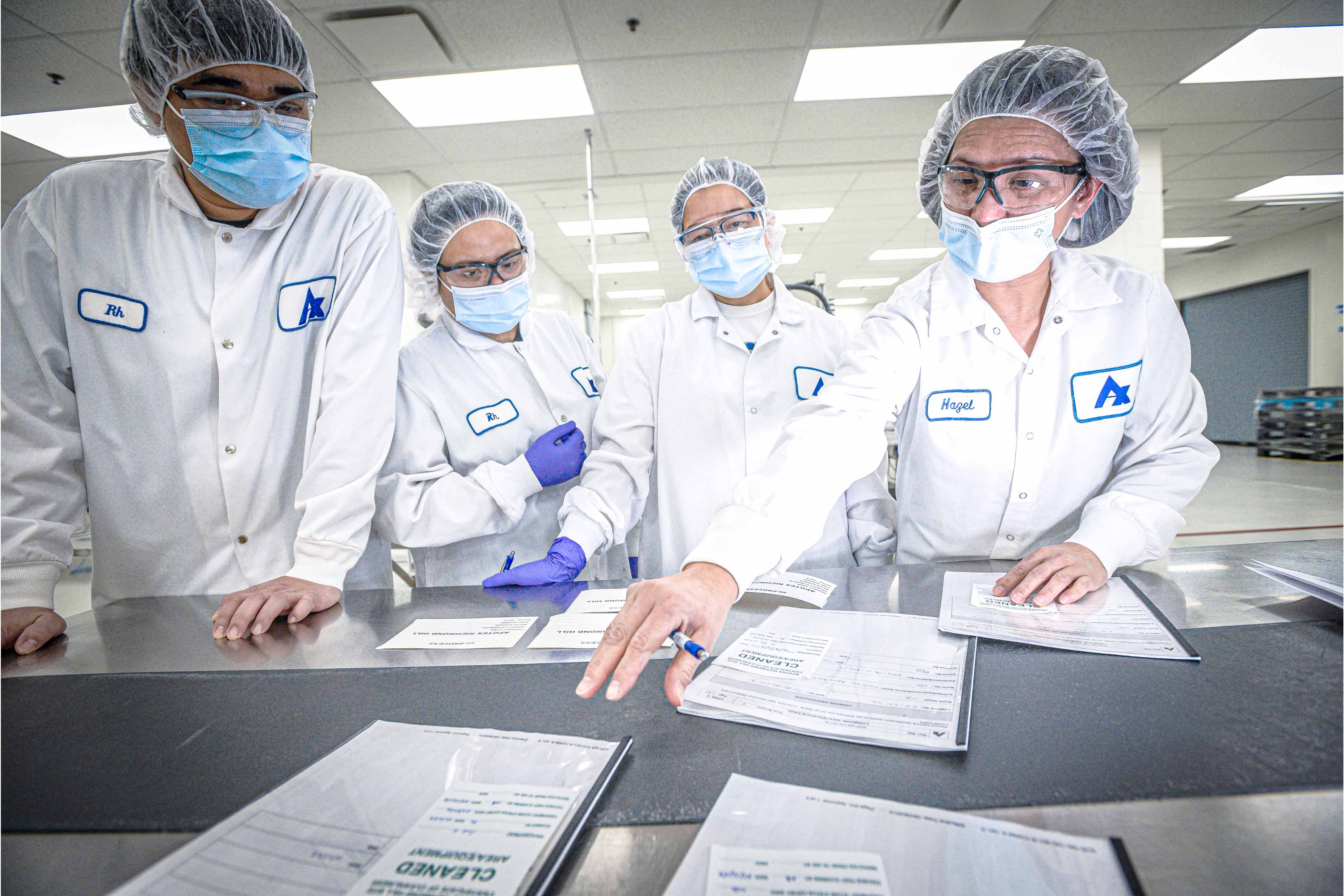 Four people in lab coats, masks, and hairnets discussing documents in a clean facility.