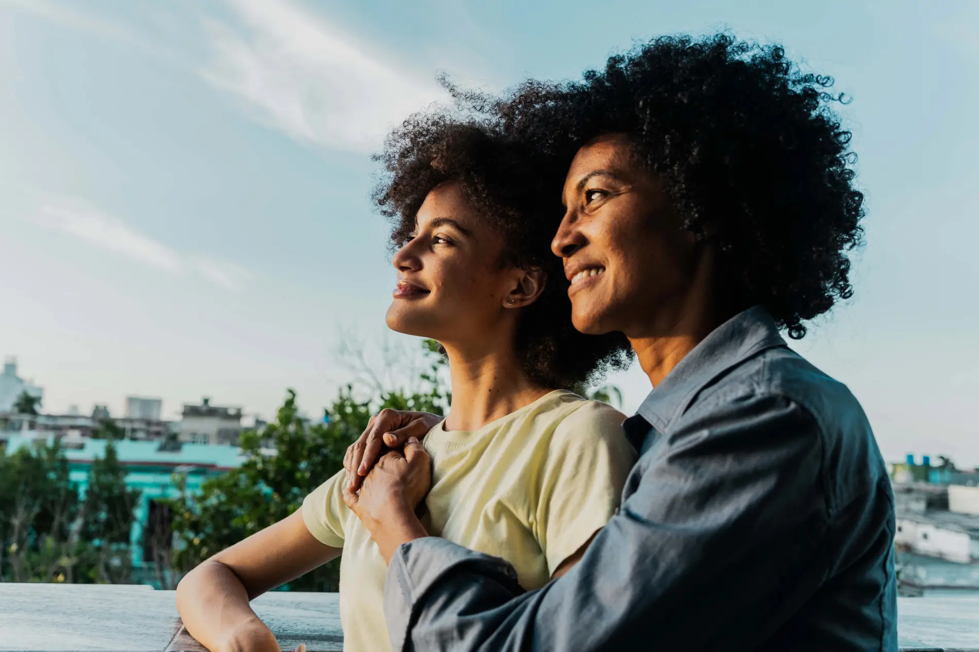 A younger woman and an older woman with curly hair stand outdoors, smiling and looking into the distance. A younger woman and an older woman with curly hair stand outdoors, smiling and looking into the distance.