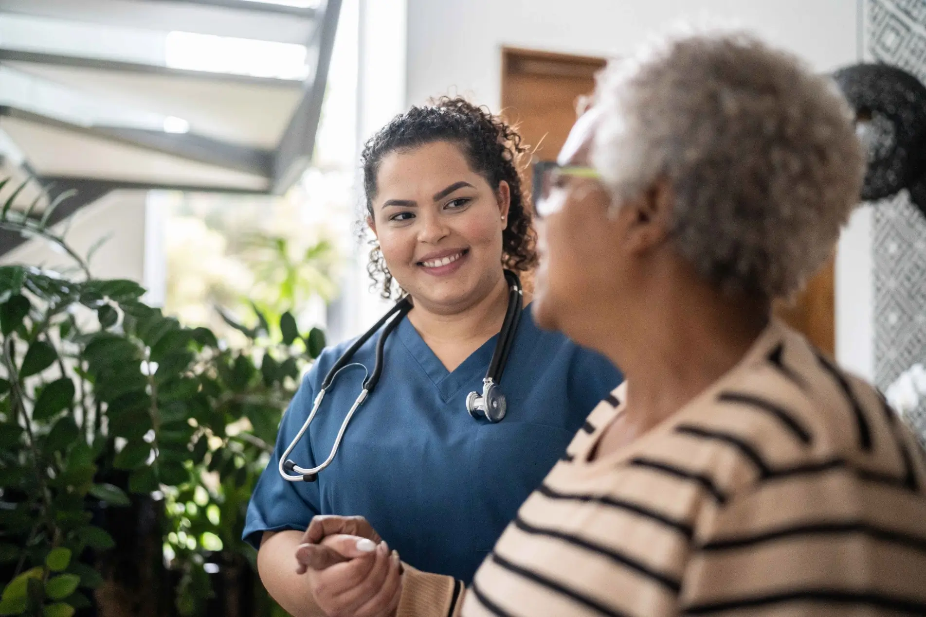 A female healthcare professional in blue scrubs smiles at an older female patient with grey hair, holding her hand.
