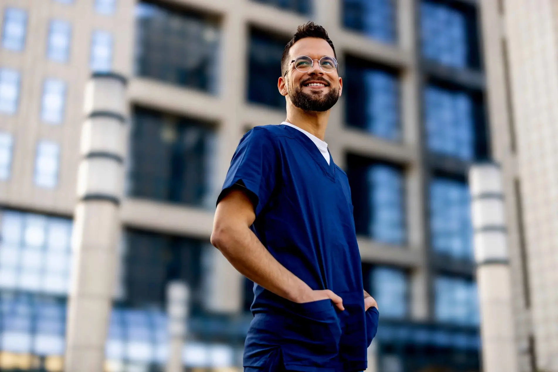 A happy man in blue scrubs and glasses standing outside a building.